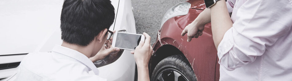 Two people looking at an auto accident, one is taking a photo of the damage that the other is pointing at
