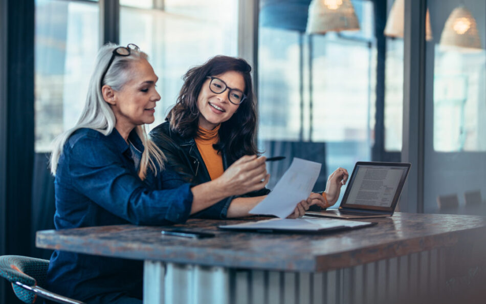 Two women analyzing documents while sitting on a table in office. Woman executives at work in office discussing some paperwork.