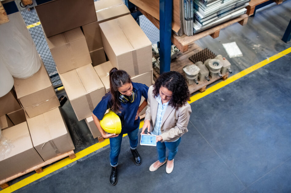 Businesswoman discussing work with female worker in warehouse. Warehouse manger showing delivery schedules on digital tablet to foreman.