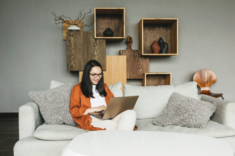 A beautiful European young woman using a laptop for remote work from home. A business woman in casual clothes conducts a video call, engages in e-mail, communicates online with colleagues, sends and responds to emails, develops software while sitting at home on the couch.
