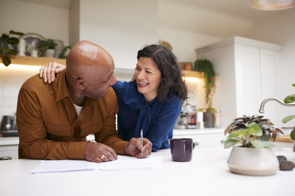 Mature Couple Reviewing And Signing Domestic Finances And Investment Paperwork In Kitchen At Home