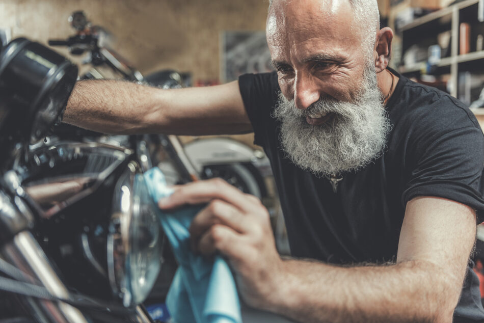 Joyous bearded mature biker is squatting near motorcycle and polishing spotlight with wide smile. He working in garage. Portrait