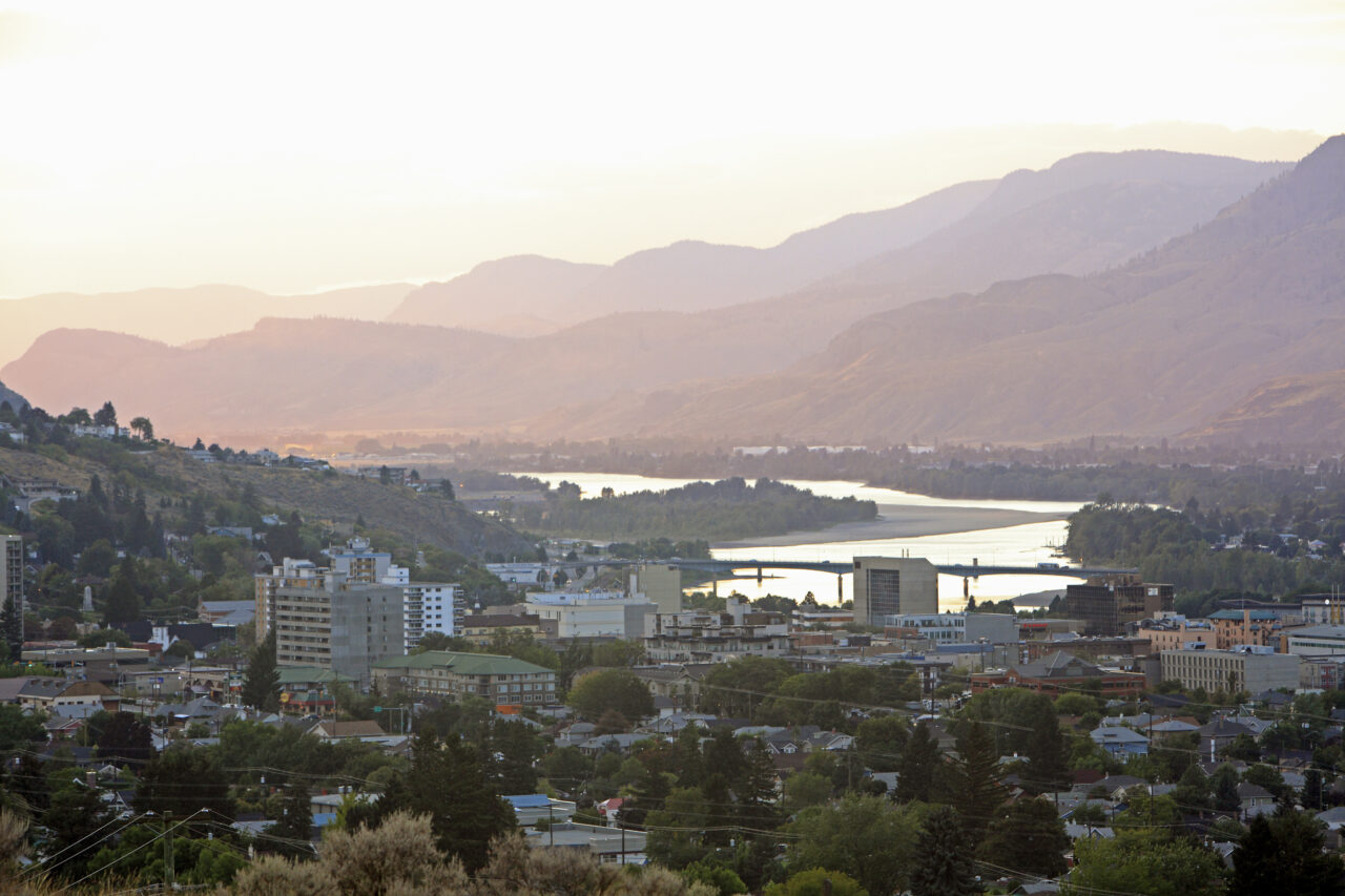 Early evening dusk over Kamloops, BC. Thompson River winds through downtown in front of layers of hills with sunlight.