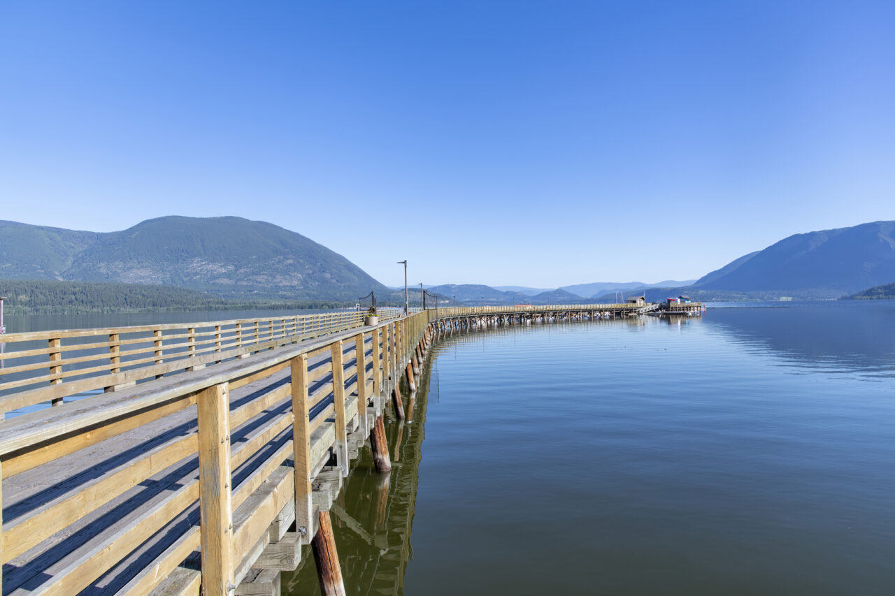 View along the curved Salmon Arm Wharf and pier with boardwalk at the shore of Shuswap Lake and panoramic view of surrounding mountains against a clear blue sky.