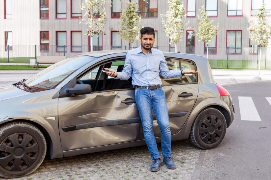 Young adult bearded man with sad expression standing with raised arms and looking at camera, being confused after car accident, broken auto with dents and scratches on side part.