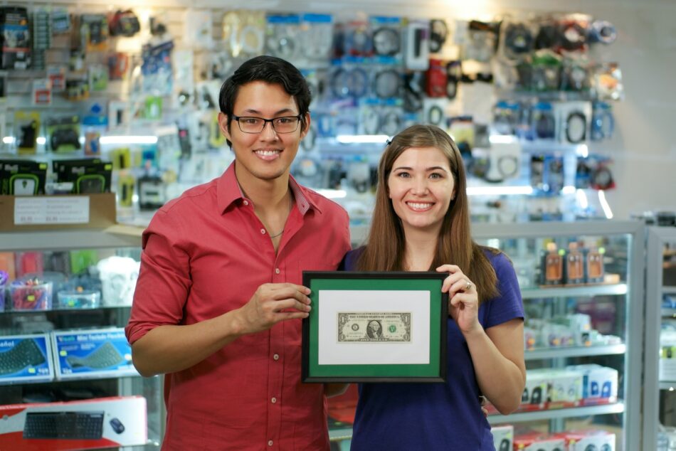 Young couple holding up a framed dollar bill and smiling in their business