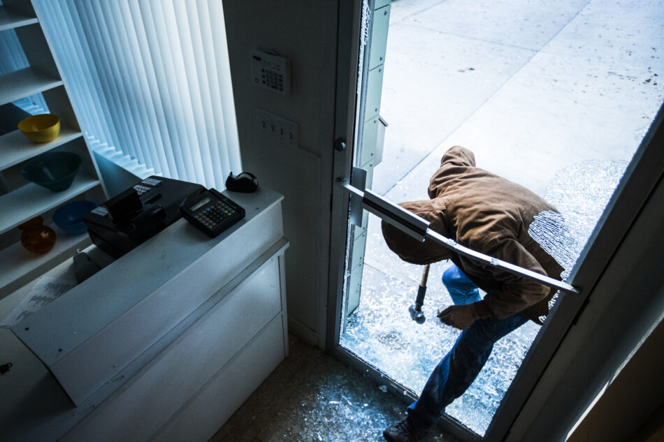 View of broken glass on the front door of a business with a hooded person performing a break and enter and ducking inside through the broken glass