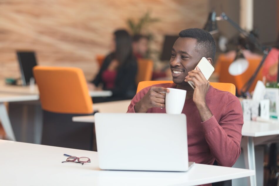 Young man sitting in a coffee shop, sipping coffee while contacting a broker on the phone with laptop open in front of him