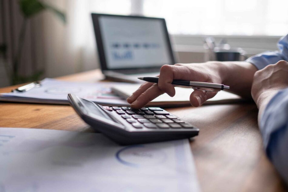 Hand pressing buttons on a calculator in front of a laptop at an office desk