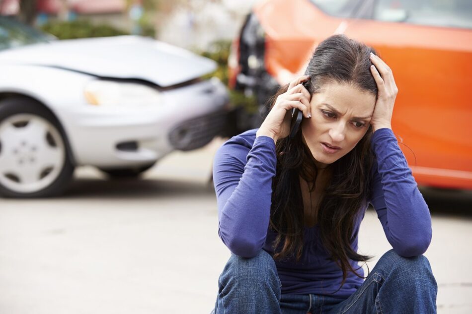 Woman sitting on curb talking on phone with fender bender behind her.