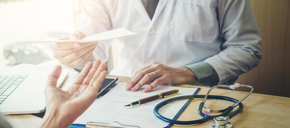 Hands at a desk with a doctor on one side talking to a patient
