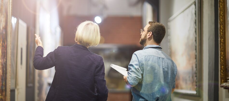 Two people in a hallway with fine art along the walls looking and discussing the art