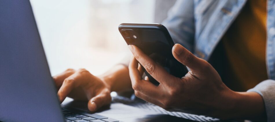 Closeup of hand holding a phone and typing on laptop at same time