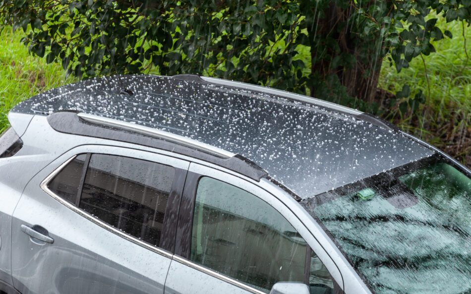 Photo of a car with visible hail damage, including dents and a cracked windshield, beneath a storm cloud with falling hail.