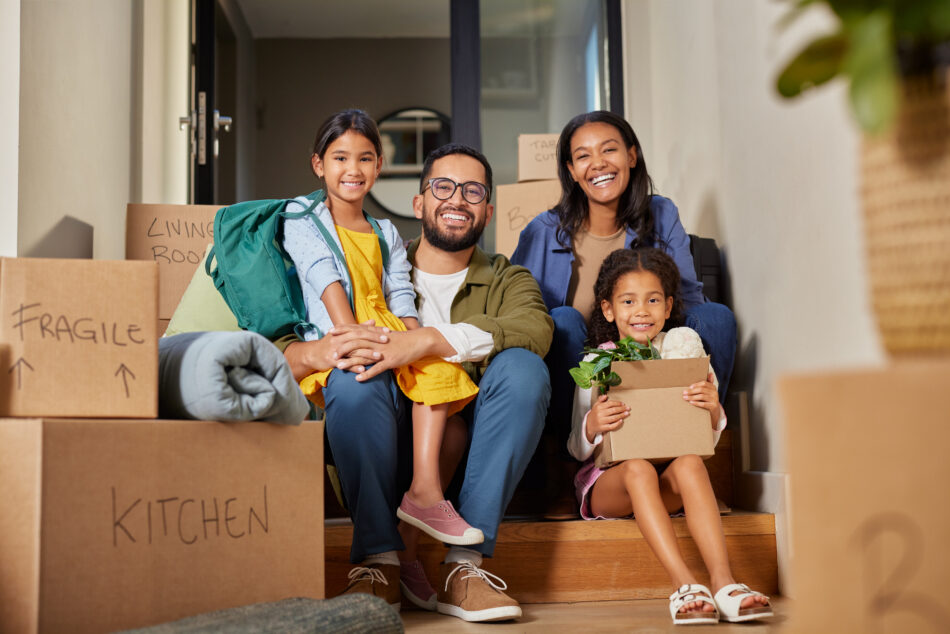 Happy young family sitting on front steps of a house and smiling at the camera while surrounded by moving boxes