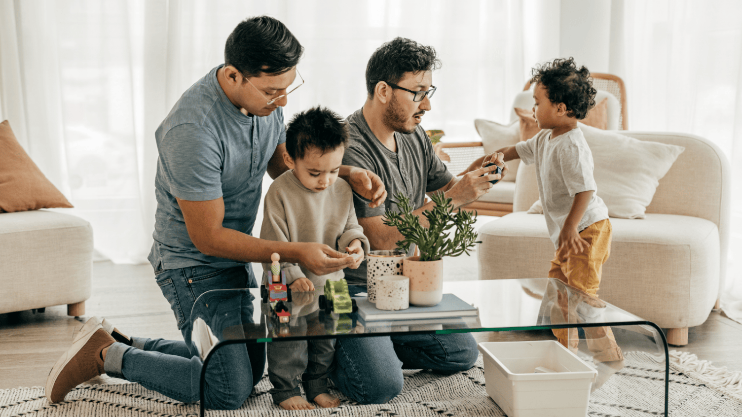 A young family of four kneel and stand around a glass coffee table in a bright living room, playing together with small toy vehicles and plants, with sofas and soft furnishings in the background.
